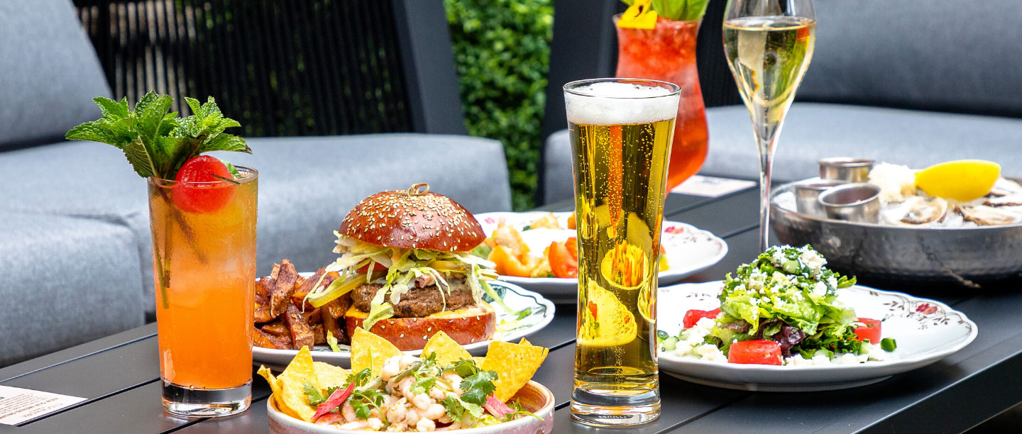 An outdoor dining table arrayed with gourmet dishes and refreshing drinks, including a tall pilsner glass of beer, colorful cocktails garnished with mint, a glass of champagne, a thick sesame-seed burger with fries, fresh ceviche with tortilla chips, a si