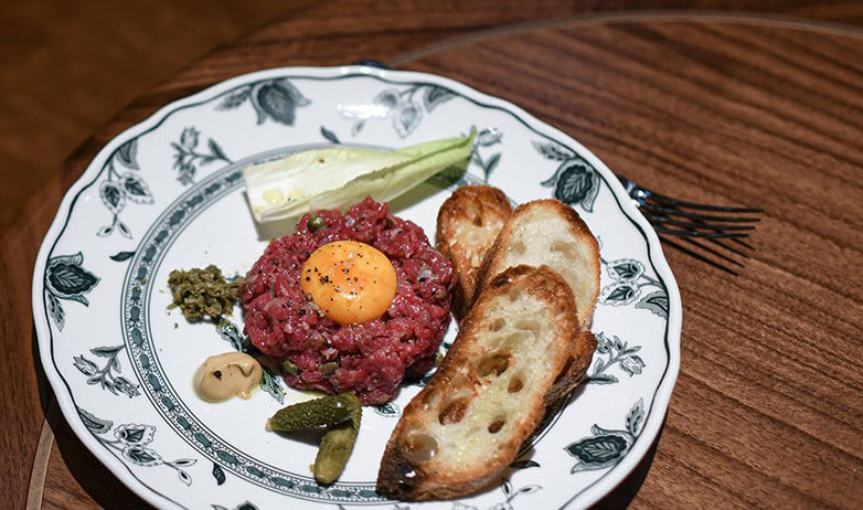 A finely plated steak tartare topped with a raw egg yolk and cracked black pepper, served on a vintage floral-patterned plate alongside toasted baguette slices, cornichons, mustard, relish, and a endive leaf.