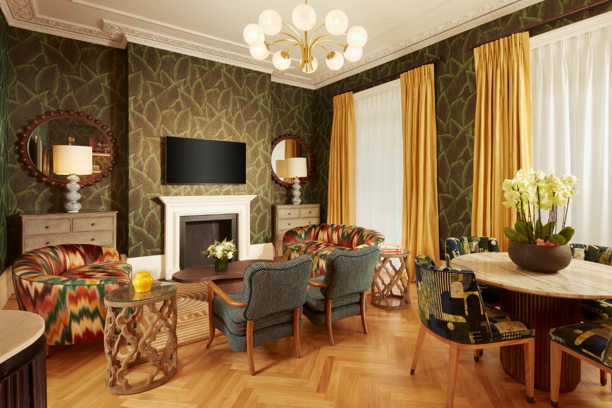 A vibrant, eclectically designed living room featuring green leaf-patterned wallpaper, colorful patterned armchairs, a white fireplace with a TV above it, yellow curtains, and a dining table.