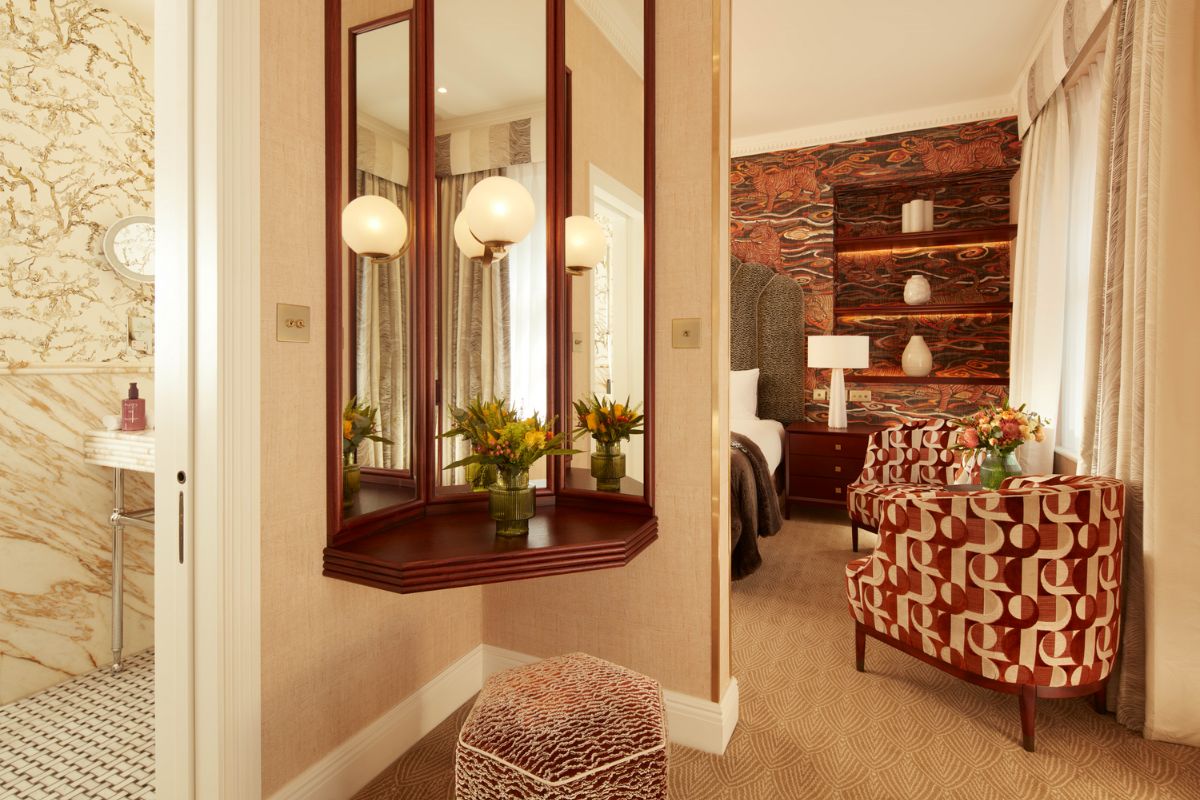 A luxury hotel bedroom entry featuring a wooden vanity with a three-panel mirror, a patterned armchair, a bed with custom tiger wallpaper, and a glimpse of the marble bathroom.