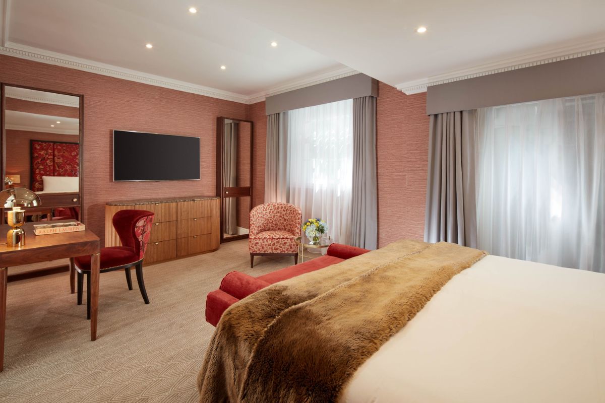 A hotel bedroom with pink textured wallpaper, a bed topped with a brown faux fur throw, a wooden desk with a red velvet chair, and a wall-mounted TV over a wooden dresser.