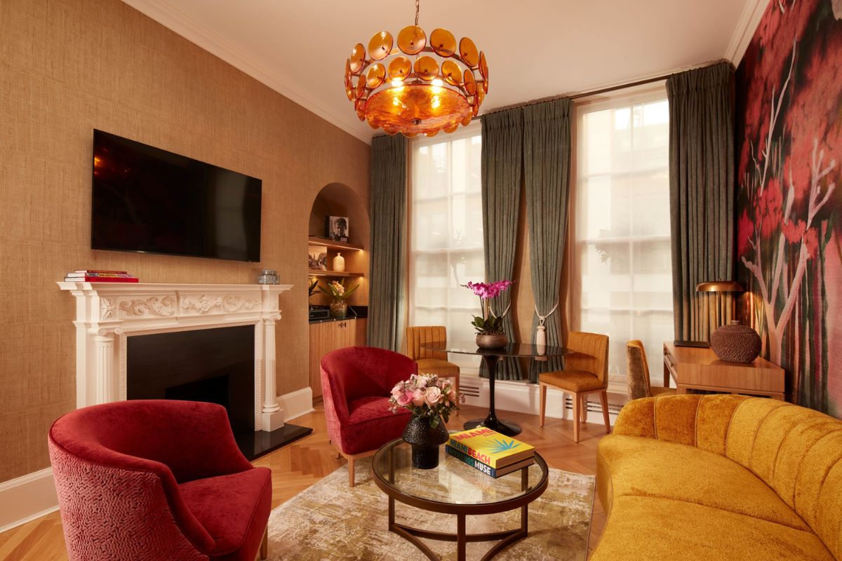 A warm, stylishly decorated sitting room with an ornate white fireplace, a yellow velvet sofa, two red armchairs, a glass coffee table, and an amber glass chandelier hanging from the ceiling.