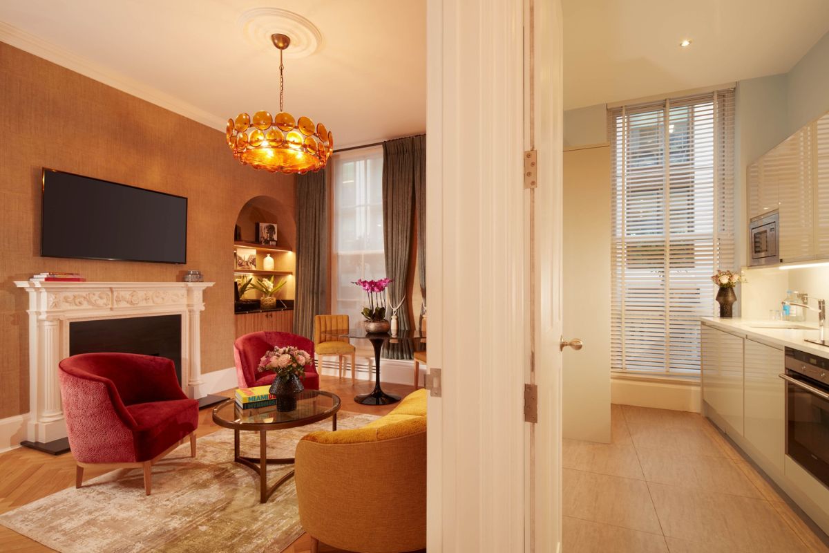 A view through a doorway showing a stylish sitting room with red armchairs, a yellow sofa, and an orange chandelier on the left, and a modern galley kitchen on the right.