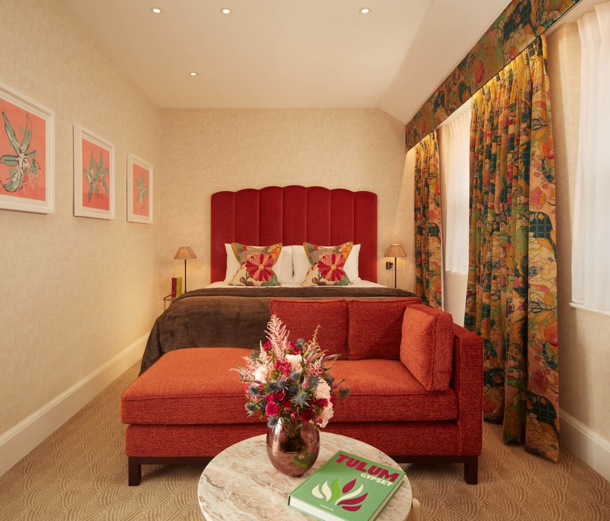 A warm hotel bedroom featuring a bed with a red scalloped headboard, a red chaise lounge sofa at the foot of the bed, a marble coffee table with a flower vase, and colorful patterned curtains.