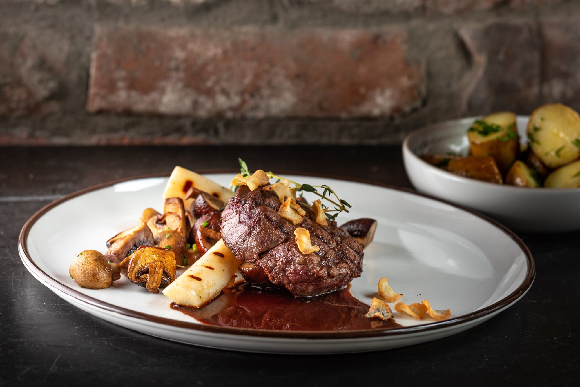 A gourmet steak dish plated on a white oval platter, garnished with crispy garlic chips, sprigs of thyme, sauted mushrooms, roasted hearts of palm, and a rich dark reduction sauce, accompanied by a side bowl of roasted potatoes.