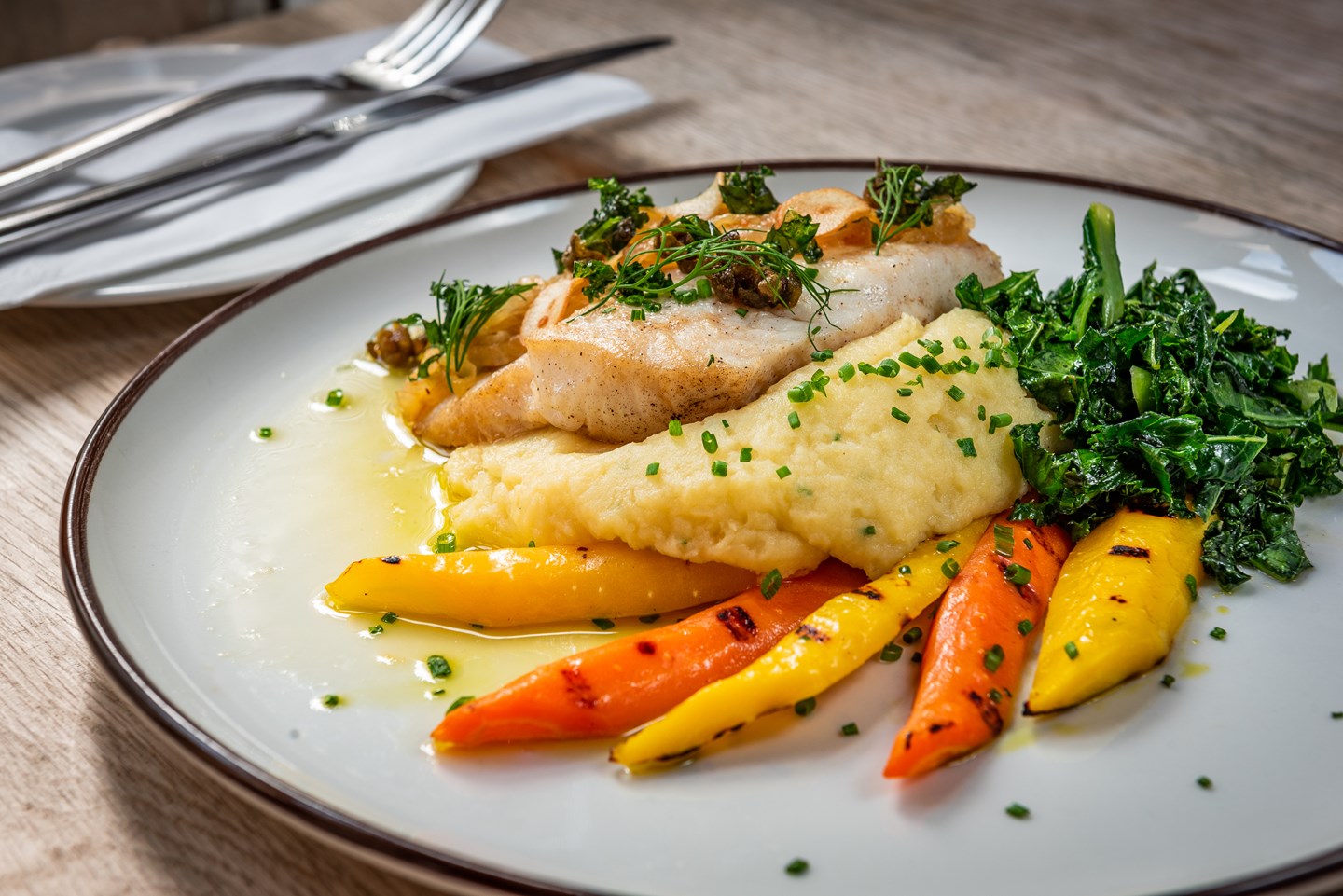 A plated seafood main course featuring a pan-seared white fish fillet topped with fresh herbs and capers, served over creamy mashed potatoes, grilled orange and yellow heirloom carrots, and sautéed dark leafy greens.