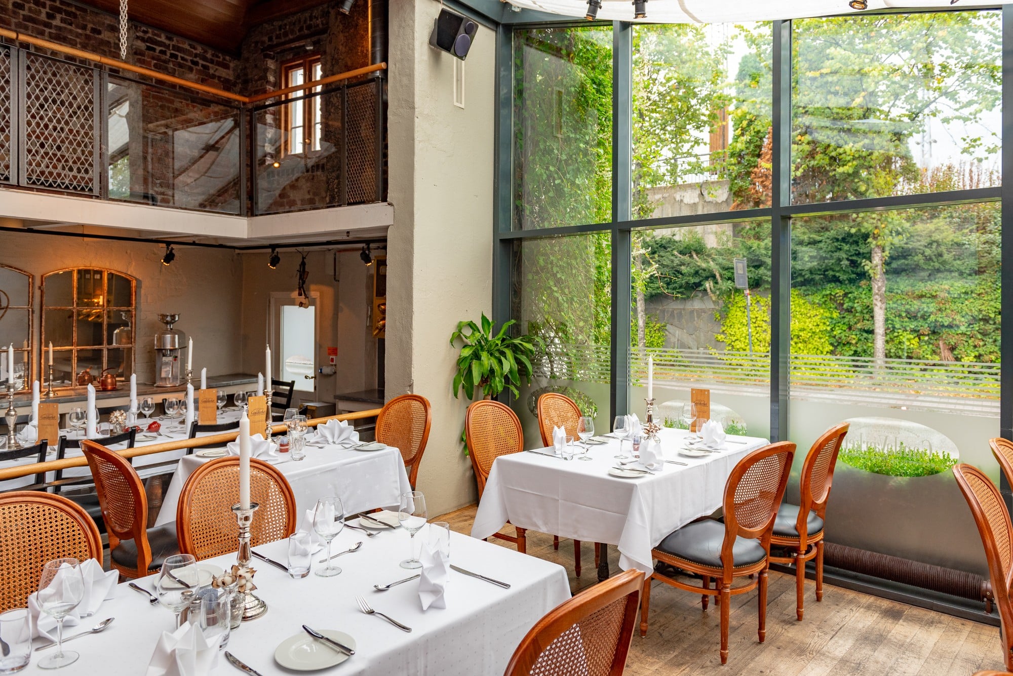 An airy, modern restaurant dining space with double-height floor-to-ceiling glass windows overlooking lush greenery, featuring white tablecloth-covered tables set with candles, wine glasses, and cane-back wooden dining chairs, alongside an exposed brick m