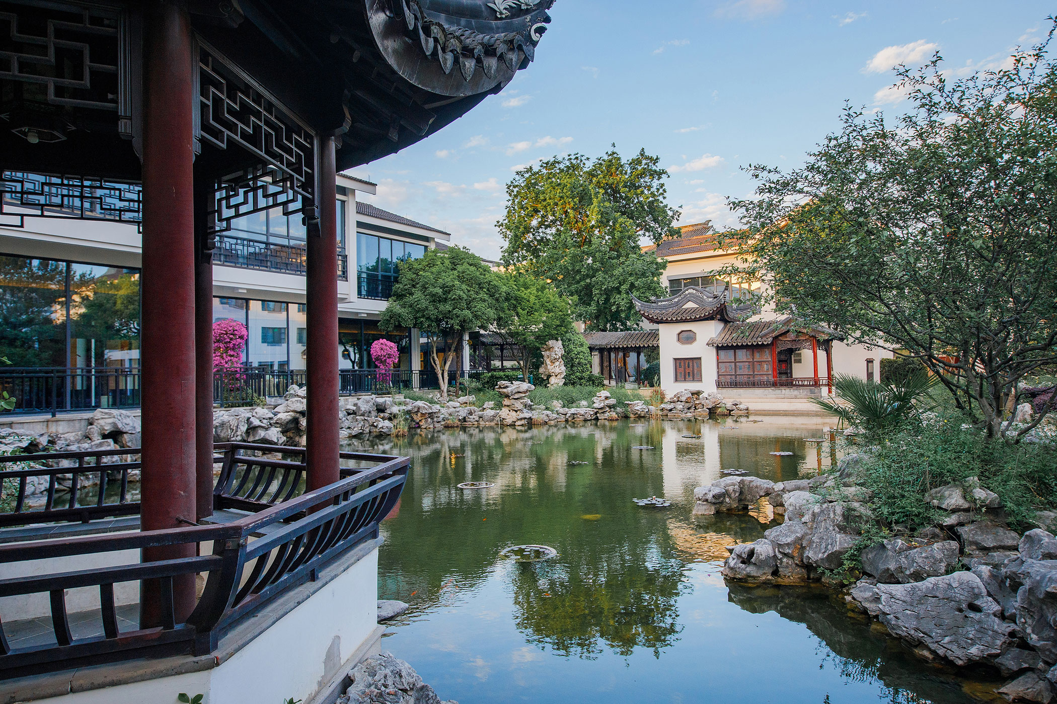 A traditional Chinese classical garden courtyard featuring a scenic pond surrounded by rocks, trees, and pavilions with distinctive curved roofs.