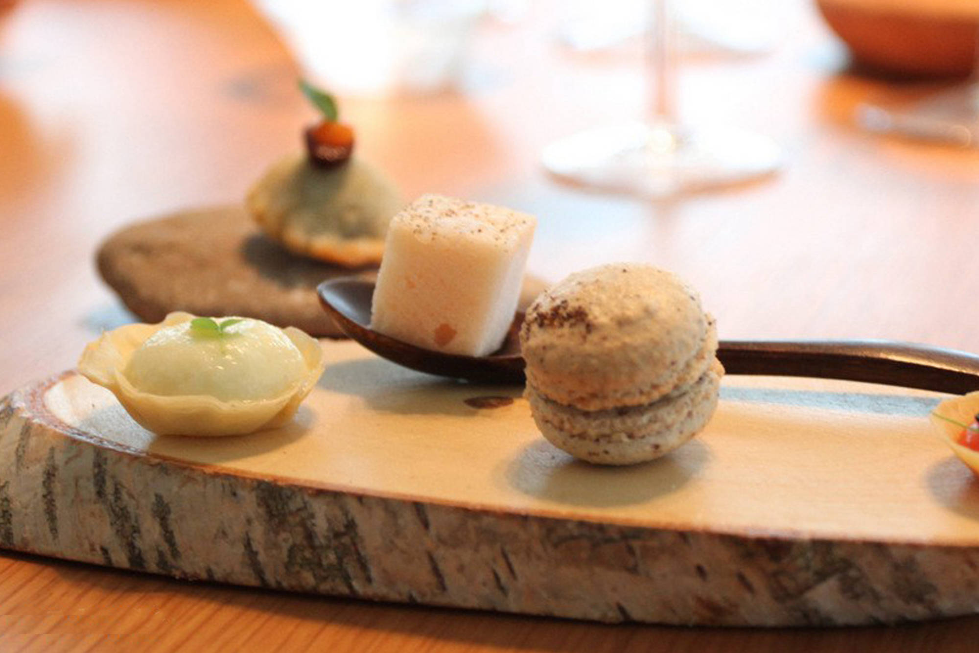 A selection of delicate, gourmet bite-sized desserts, including a macaron and a marshmallow cube on a spoon, elegantly served on a wooden log slab.