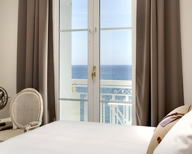 A view from a bedroom looking through white glass doors out to a balcony and the blue ocean, framed by brown curtains.