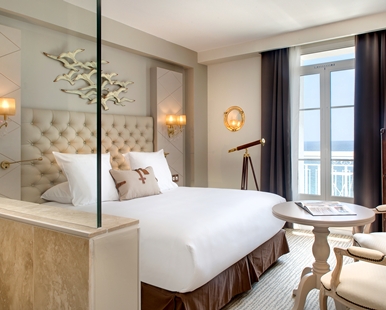 A nautical-themed hotel room featuring a bed with a beige tufted headboard, a brass decorative telescope, bird wall art, and glass doors looking out to a balcony and the sea.