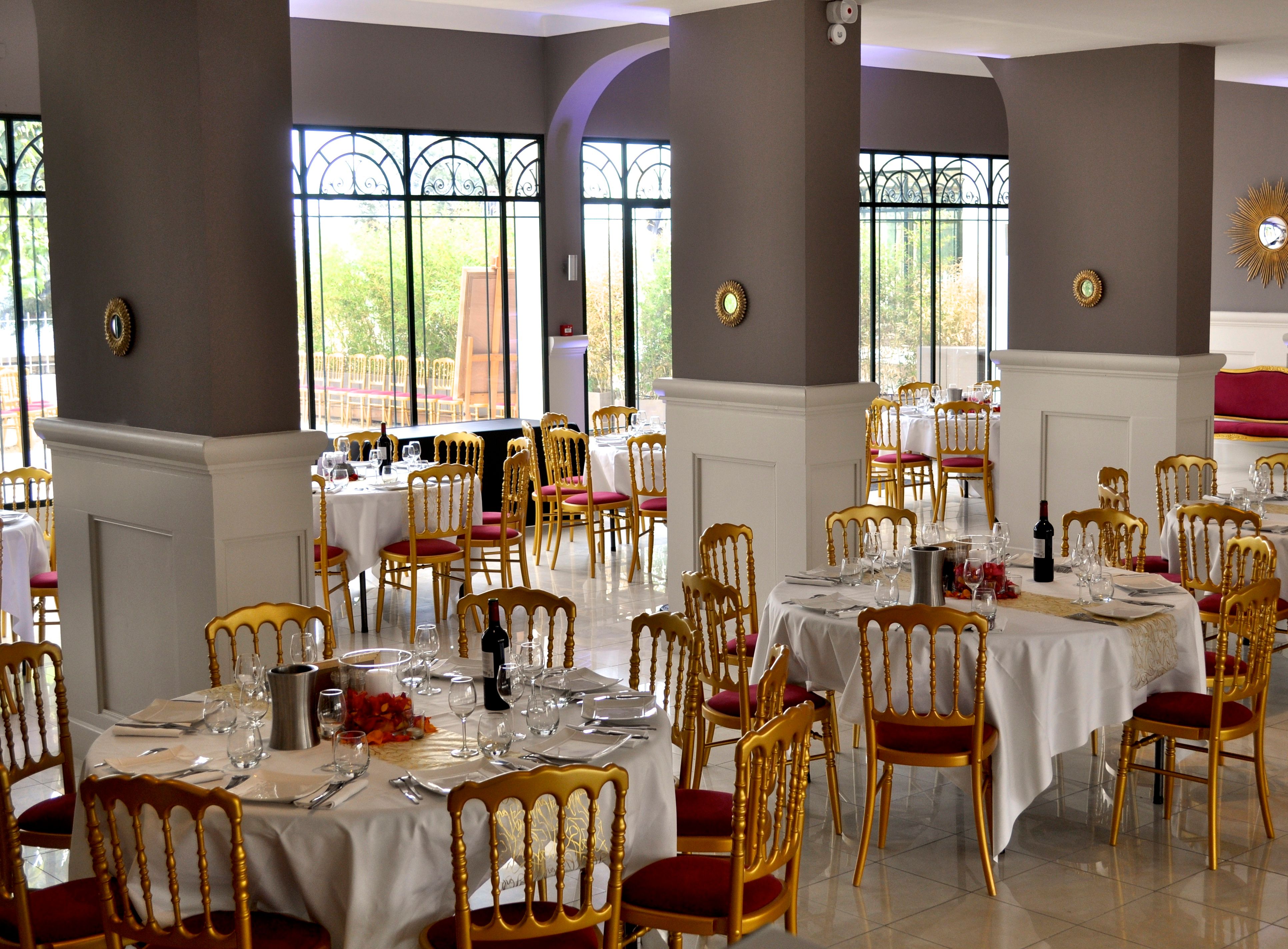 A brightly lit event dining room set up with multiple round tables, white linens, wine bottles, and elegant gold-framed chairs with red cushions.