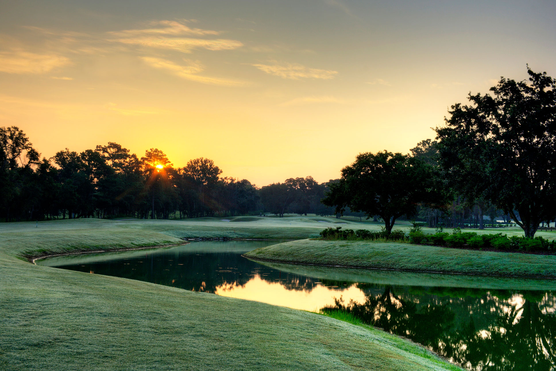 A scenic golf course fairway with a winding water hazard reflecting trees and a bright golden sunrise.