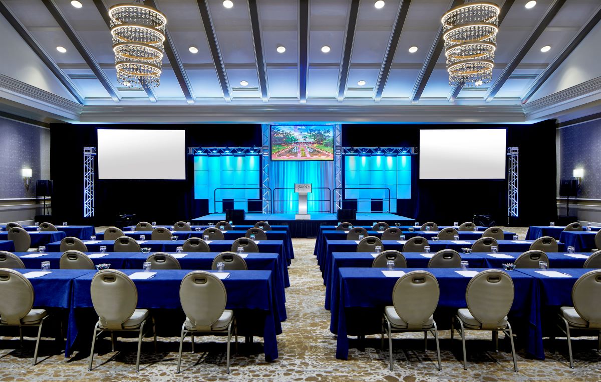 Large conference room set up with blue-clothed tables, chairs, two spiral chandeliers, and a stage with presentation screens.