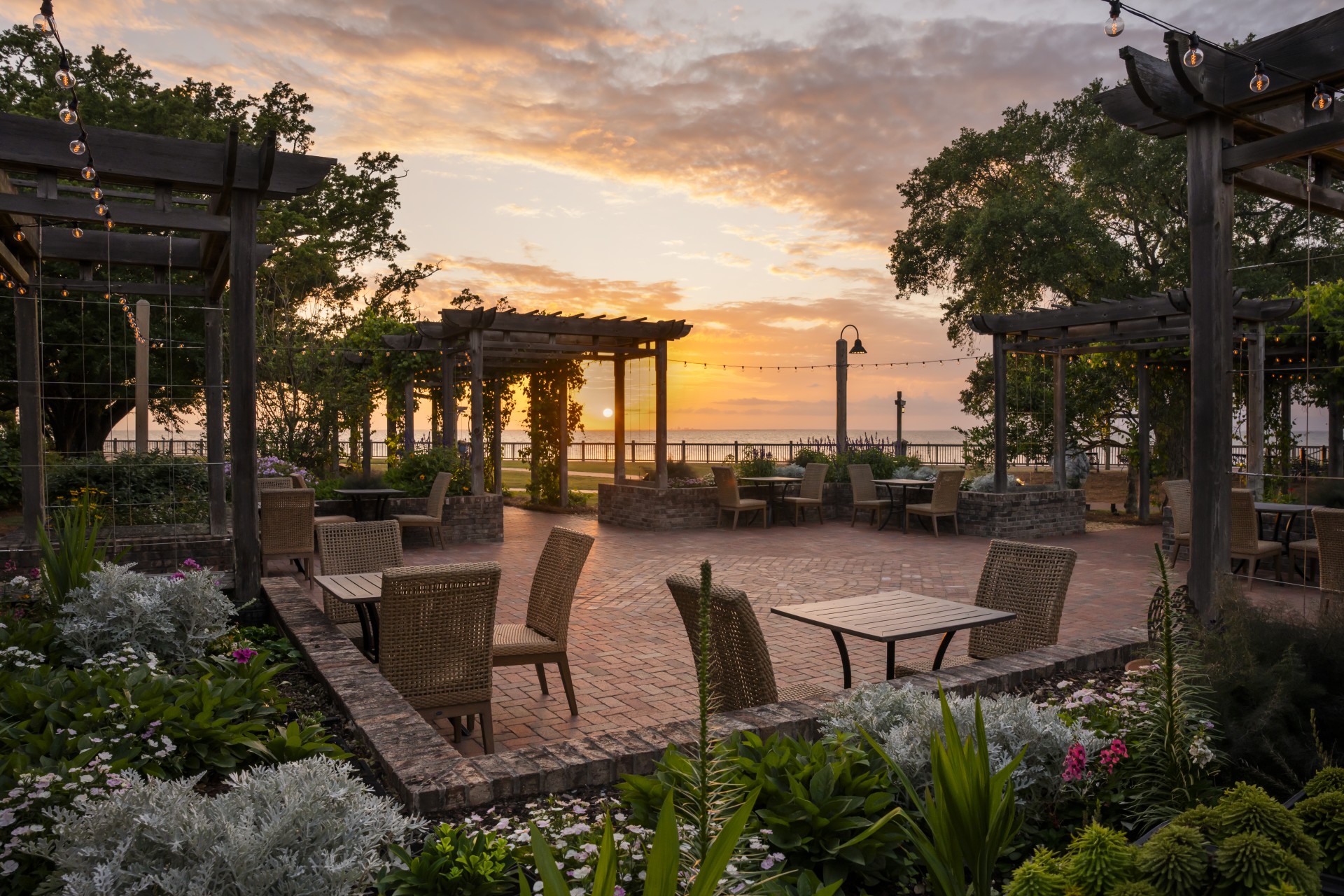 An outdoor brick patio featuring wooden pergolas, wicker patio seating, surrounding flower beds, and a scenic sunset view over the water.