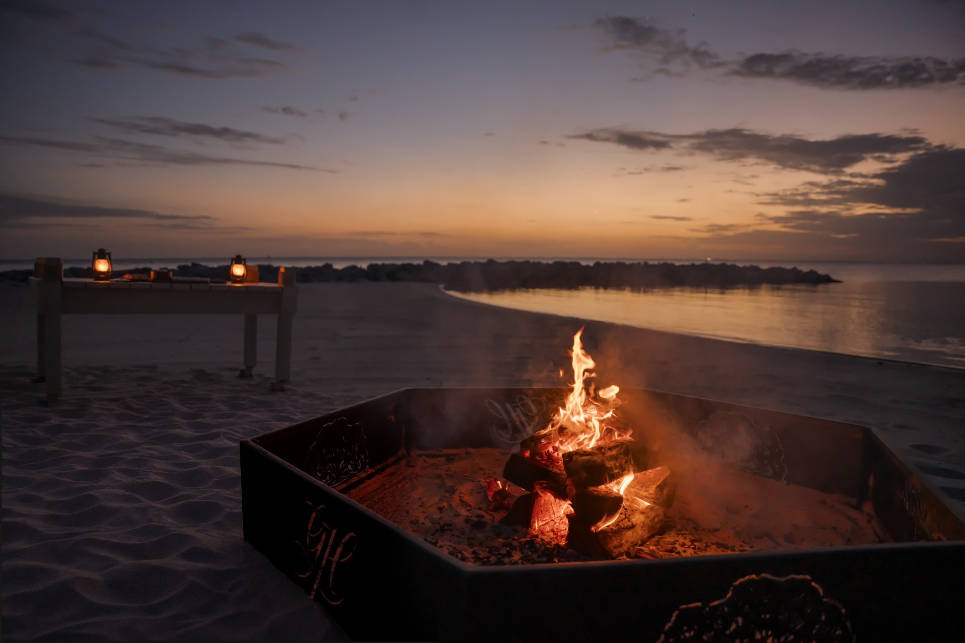 A crackling fire pit on a sandy beach at twilight, with lanterns lit on a table near the water's edge.