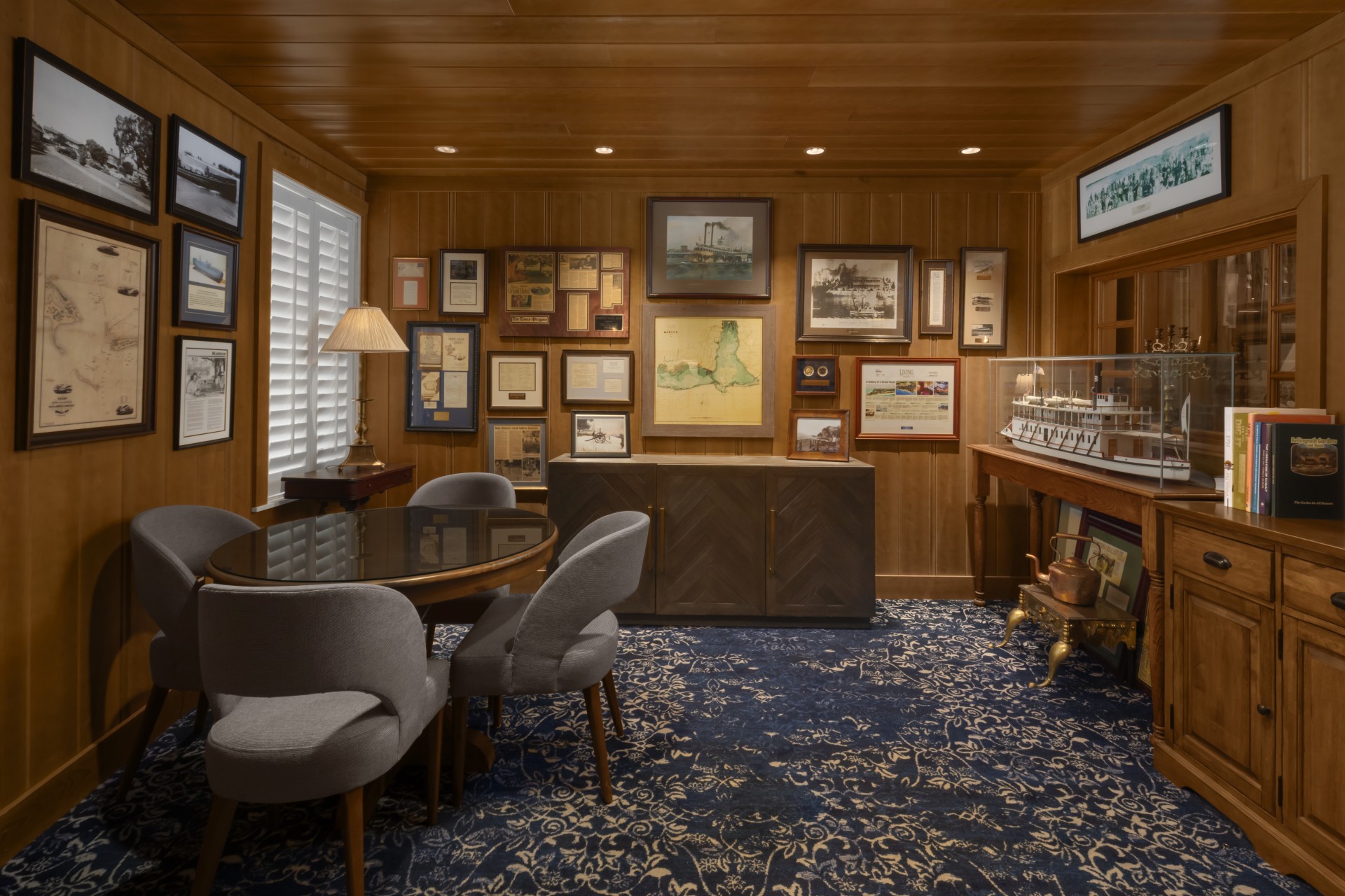 Cozy, wood-paneled lounge with framed historical memorabilia on the walls, a small round meeting table with grey chairs, and a scale model ship.