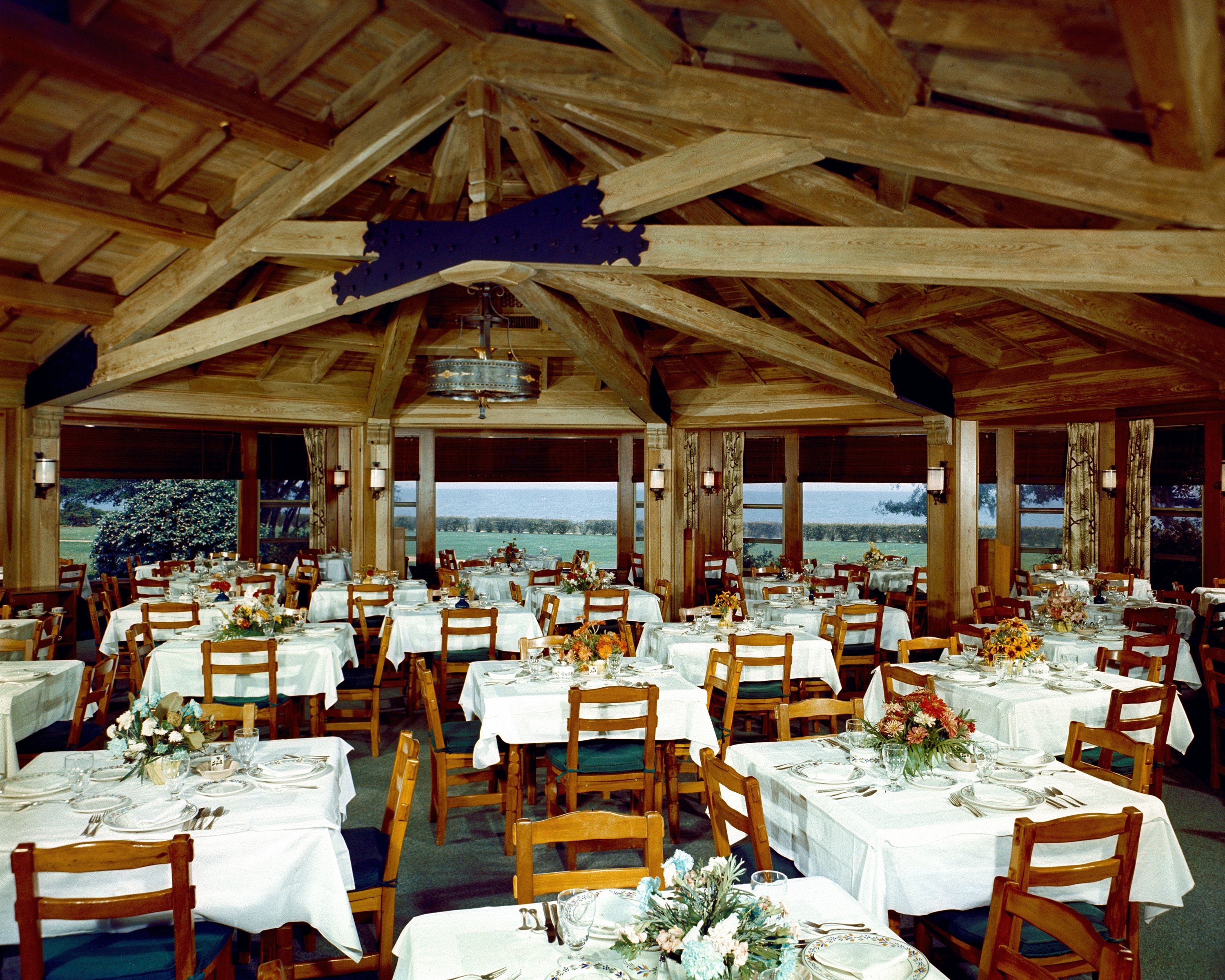 Rustic dining hall with exposed wooden ceiling beams and tables set with white tablecloths overlooking a grassy landscape.