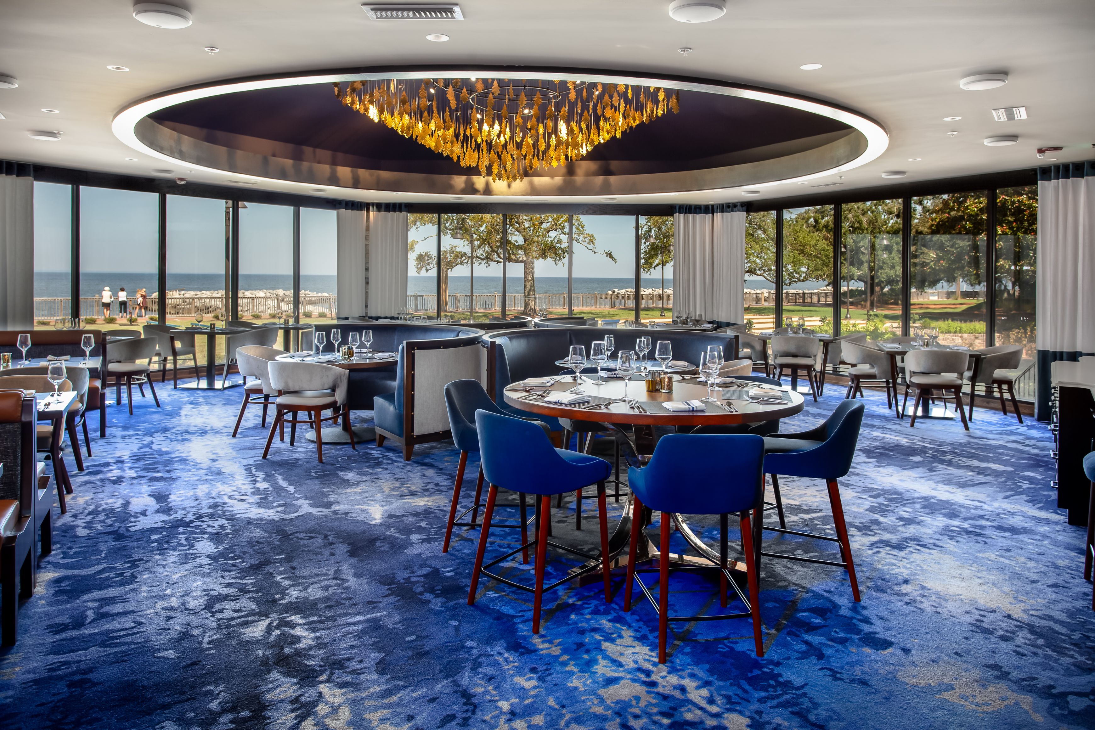 Modern upscale restaurant dining room with blue carpet, gold chandelier, and floor-to-ceiling windows overlooking the ocean.