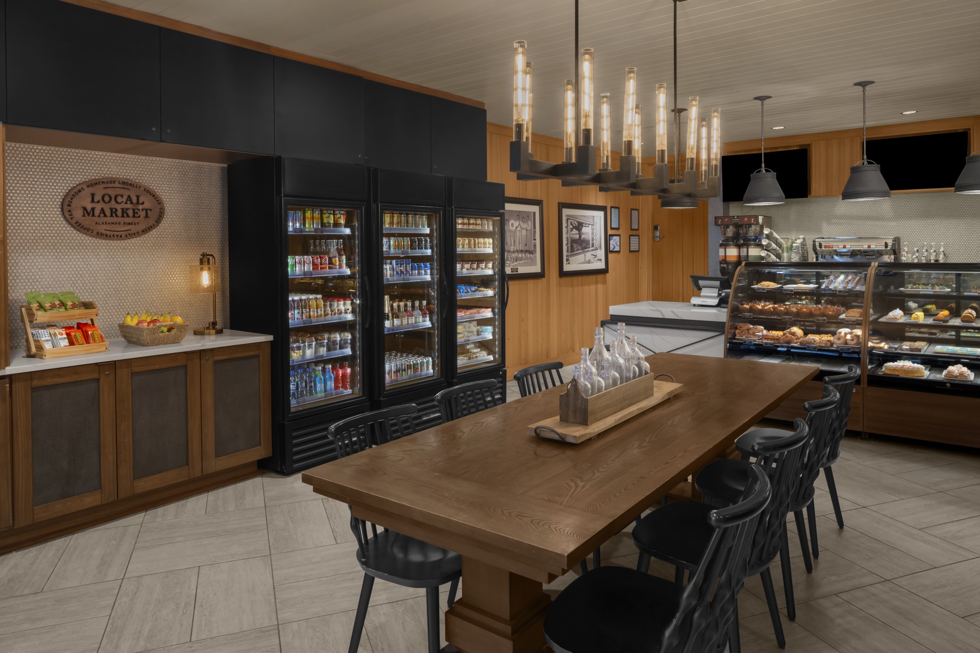Hotel grab-and-go local market area featuring beverage coolers, pastry display cases, and a wooden communal dining table.