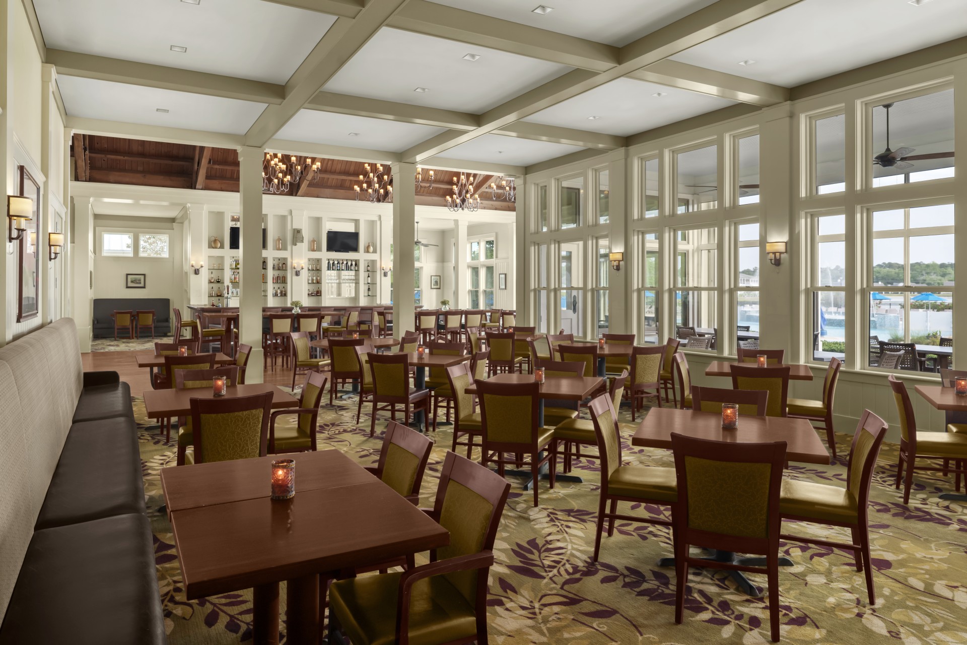 Bright resort dining room with coffered ceilings, large windows, wooden tables, and a bar area in the background.