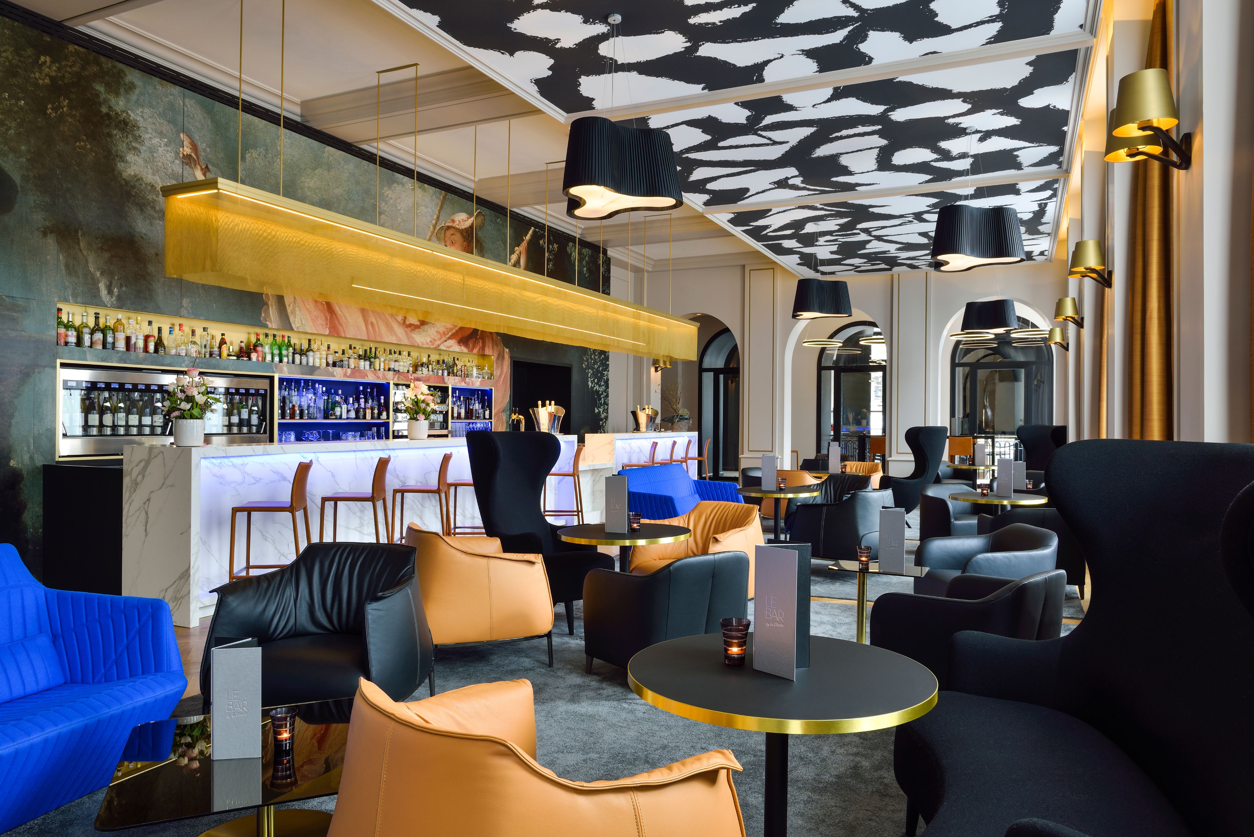 A stylish modern hotel bar and lounge featuring an illuminated marble bar counter beneath a long gold mesh light fixture, paired with vibrant blue, tan, and black leather armchairs, black small round tables, and a bold black-and-white patterned ceiling.
