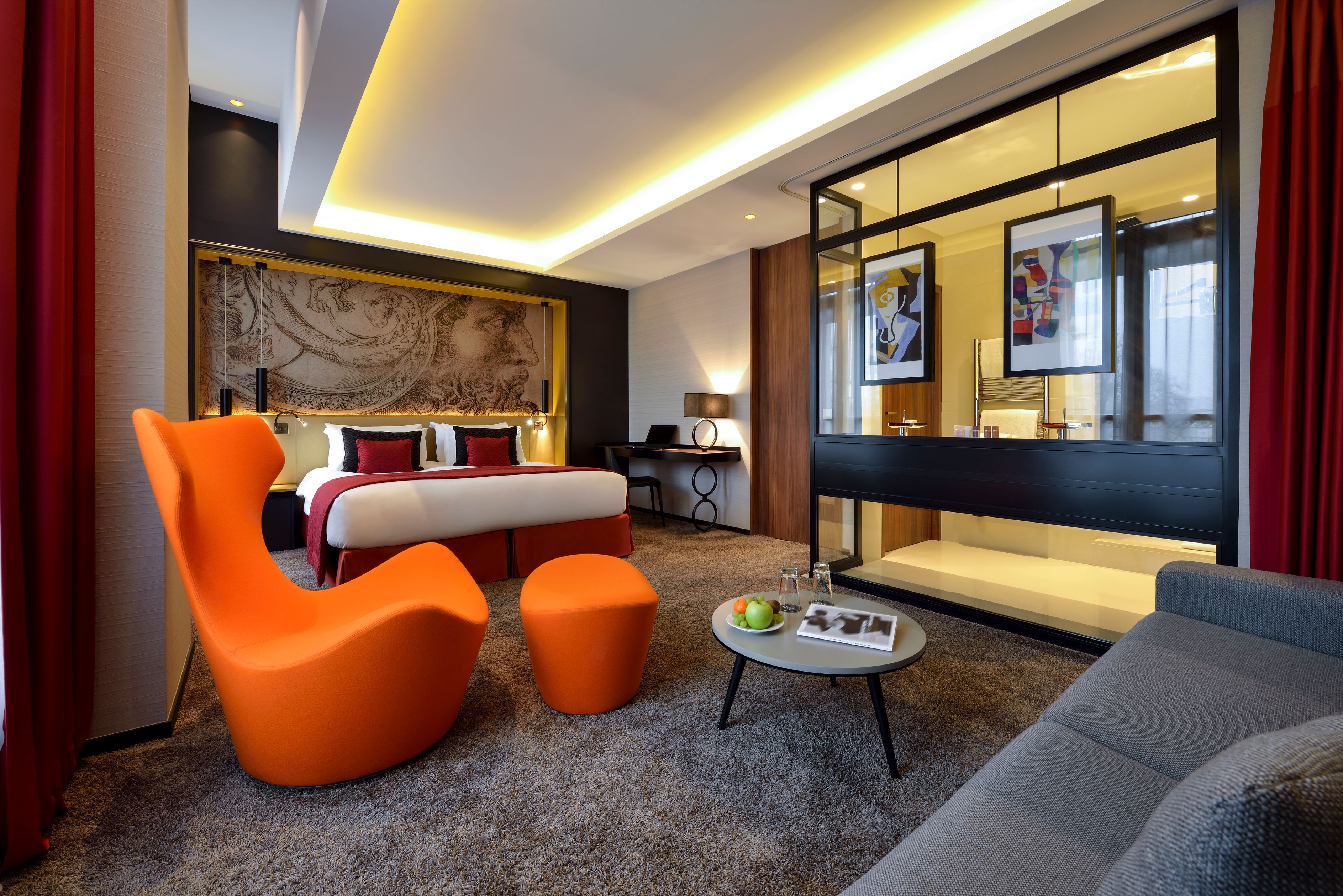 A modern, chic hotel suite featuring a bright orange lounge chair and ottoman in the foreground, a king-sized bed with a dark classic mural accent wall, warm recessed ceiling lighting, and a glass partition separating the bedroom from an open-concept bath