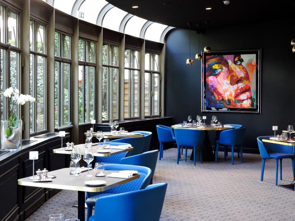 A contemporary dining space with royal blue tub chairs, dark walls, a glass solarium-style window wall, and a large colorful abstract portrait painting on the back wall.