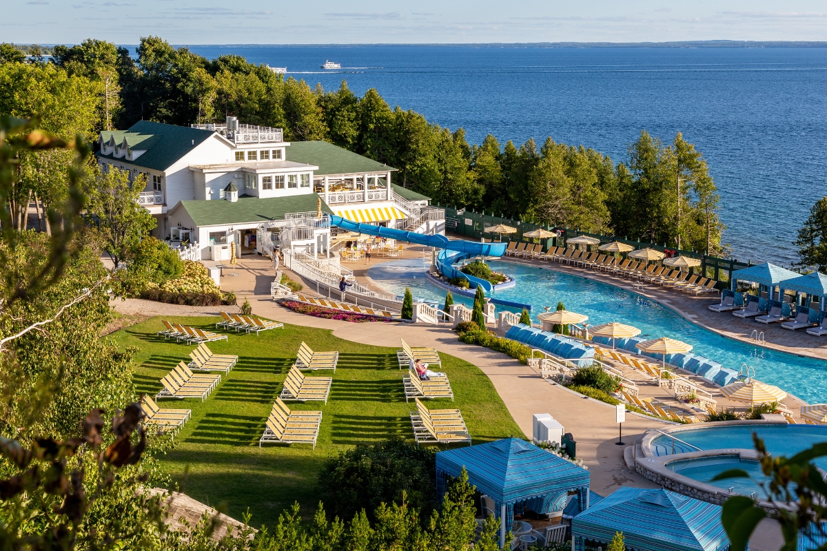 Aerial view of a lakeside resort featuring a large swimming pool complex with a blue waterslide, sun loungers spread across a grassy lawn, blue poolside cabanas, and a white multi-level building surrounded by trees.