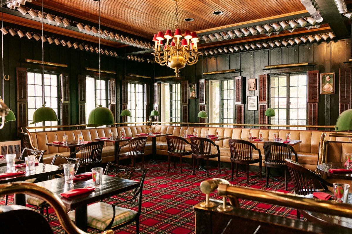 Equestrian-themed dining room featuring red tartan carpet, tan leather banquettes, green riding helmet lights, a red-shaded chandelier, and pewter mugs lining the ceiling beams.