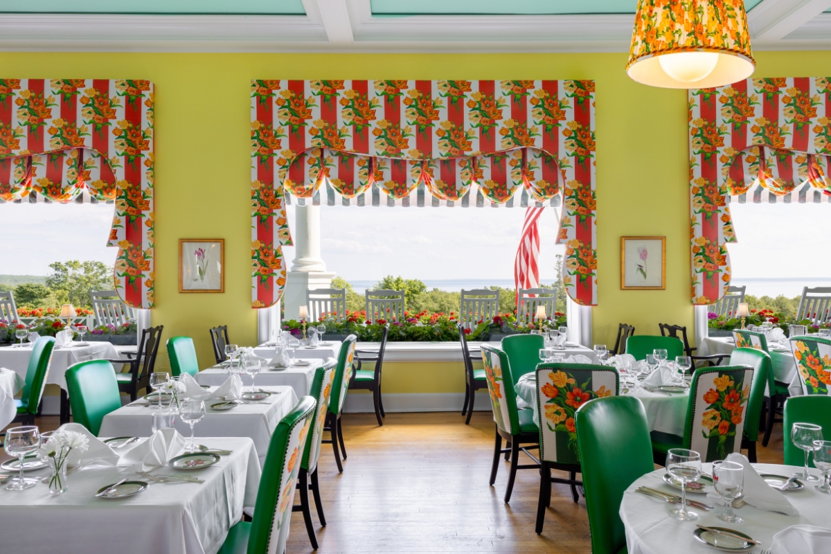 Bright dining room featuring yellow walls, light blue ceiling, green upholstered chairs with floral-patterned backs, matching red-and-yellow floral valances, and large windows looking out onto a porch and water view.