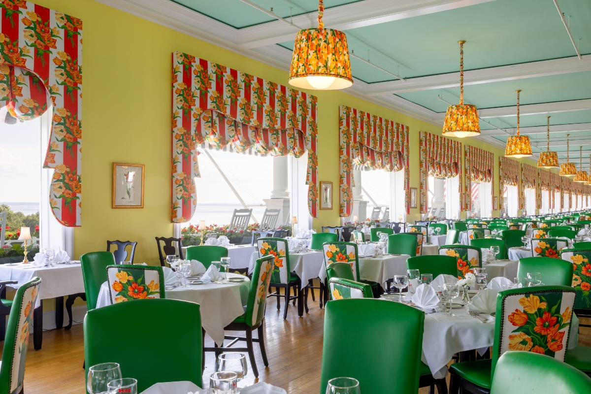 Vibrant restaurant dining room with yellow walls, a light blue ceiling, green dining chairs with floral-patterned backs, matching floral valances, and hanging floral lampshades.