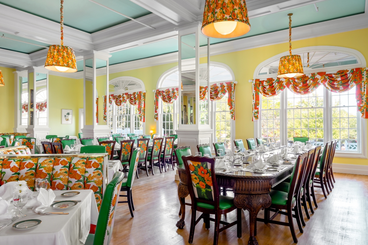 Spacious dining hall with soft yellow walls, sky-blue tray ceiling, floral pendant lights, large arched windows, and dark wood tables surrounded by green chairs featuring vibrant orange-and-yellow floral upholstery.