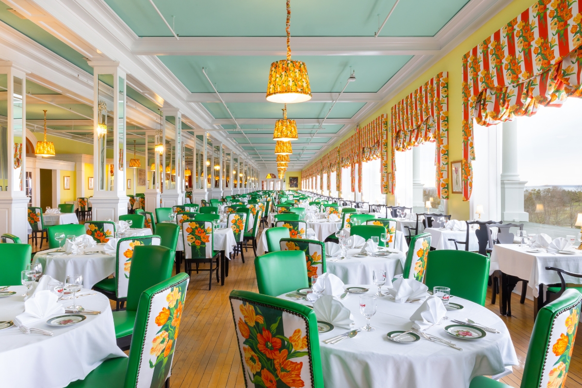 Grand dining room filled with white-cloth tables, green chairs featuring floral backs, hanging floral lampshades, yellow walls, and a mint-green ceiling.