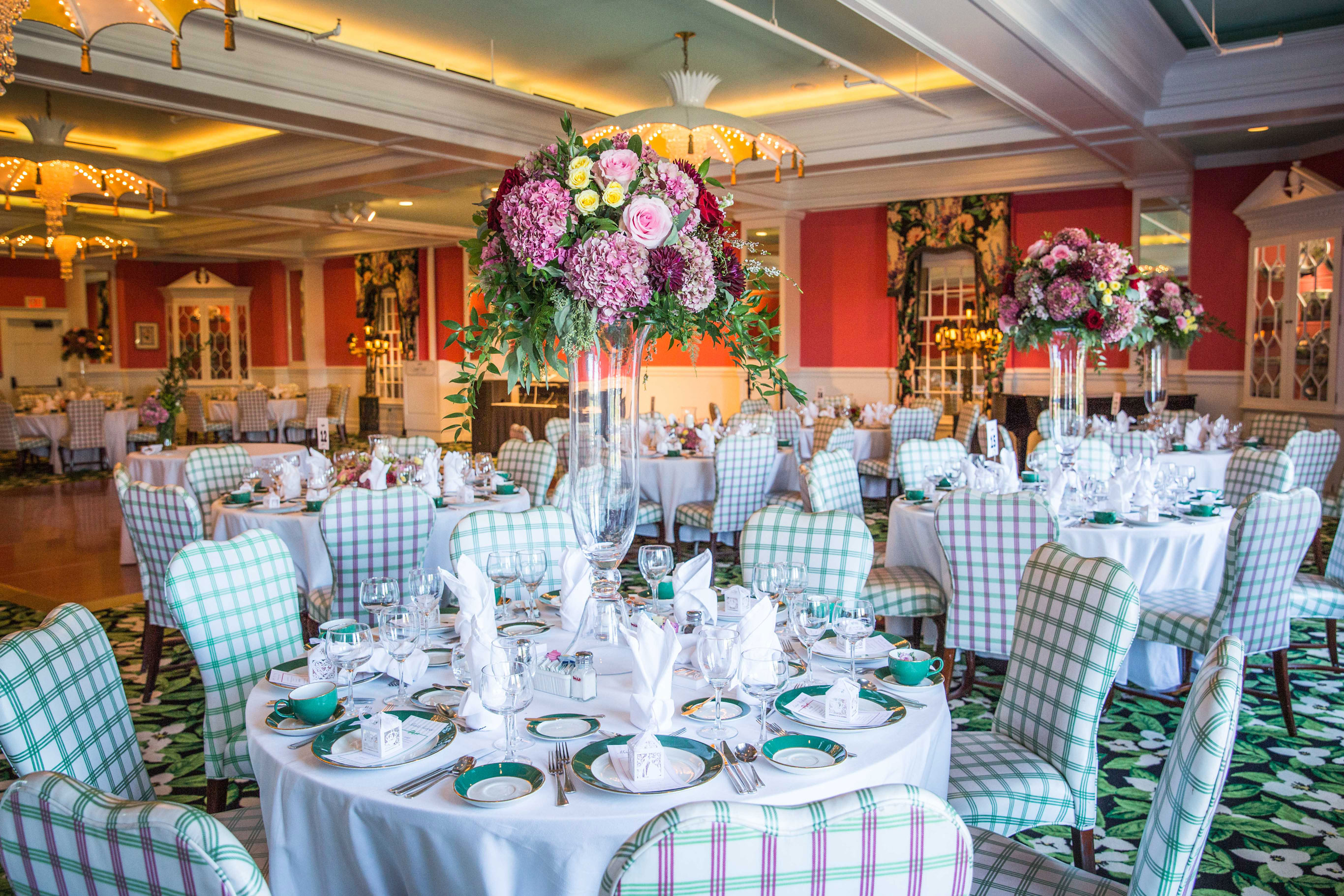 Elegantly set event room with round tables, green and white plaid chairs, tall floral centerpieces featuring pink and purple flowers, coral walls, and decorative umbrella-style chandeliers.