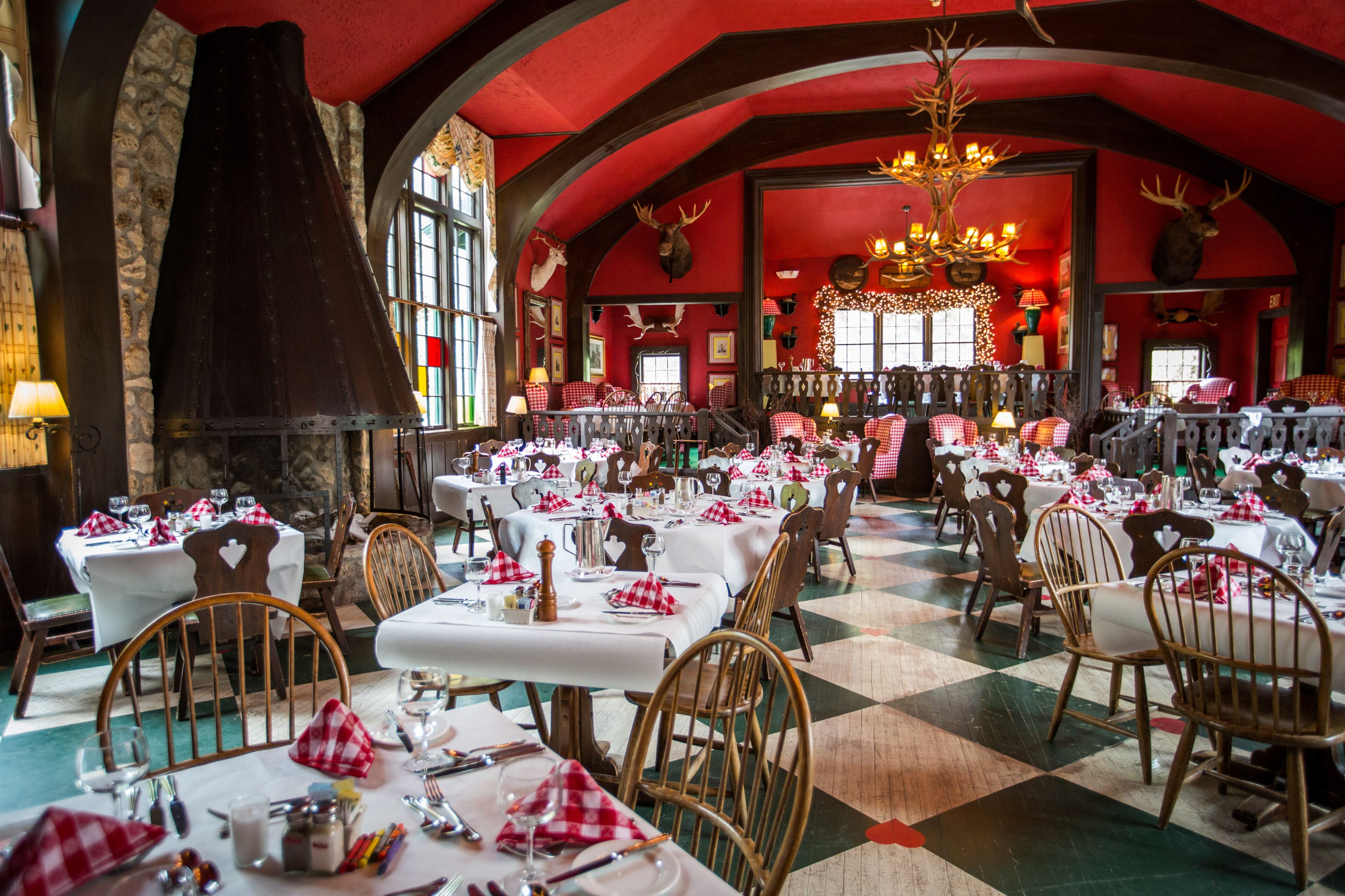 Rustic lodge dining room with a vibrant red ceiling, dark wood ceiling beams, antler chandeliers, taxidermy mounts, a conical stone fireplace, and diamond-patterned flooring.