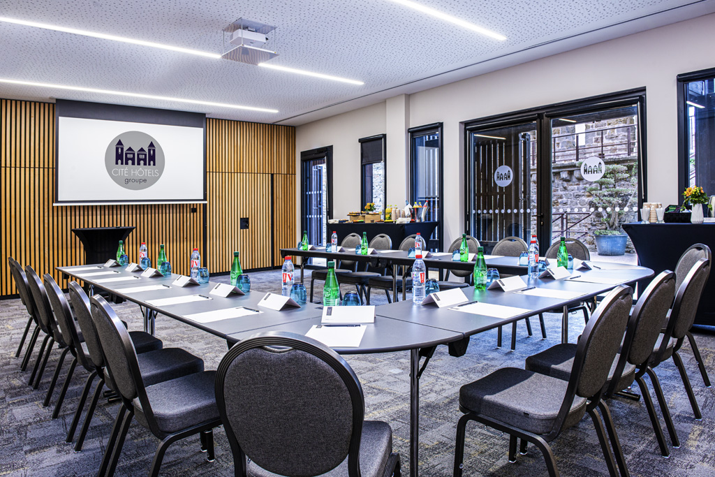 A modern, brightly lit conference room arranged in a U-shape layout with grey padded chairs and dark tables, featuring a wall-mounted projection screen displaying the 'Cité Hôtels groupe' logo, vertical wood slat wall accents, and glass doors open