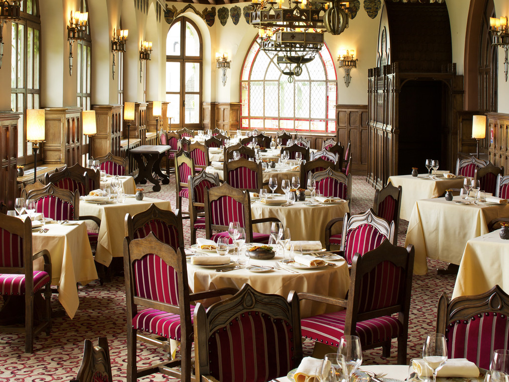 A grand gothic-style dining hall set up with formal table arrangements, featuring dark carved wooden chairs with red and gold striped upholstery, cream tablecloths, iron chandeliers, wainscoting, and large arched leaded glass windows.