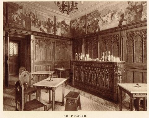 A historical sepia-toned photograph labeled 'LE FUMOIR', showing a gothic-style smoking lounge or bar with carved wood wainscoting, an ornate wooden bar counter with liquor bottles, small tables with wooden chairs, and a detailed mural frieze runn