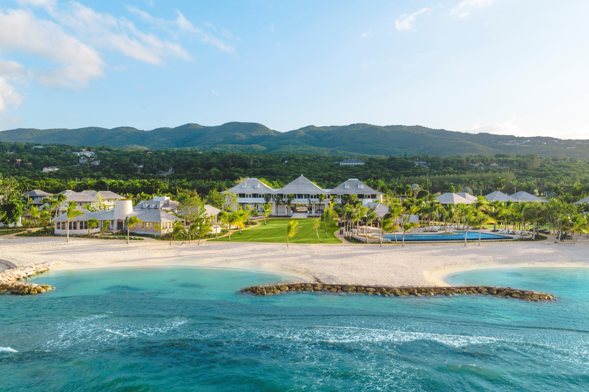 A wide aerial view of a beachfront luxury resort with white sand, turquoise ocean water, palm trees, a sprawling green lawn, outdoor swimming pool, and lush green mountains in the background.