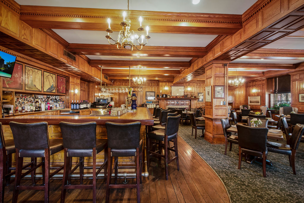Cozy, wood-paneled pub and dining area with a classic wooden bar counter, black leather barstools, brass chandeliers, and adjacent carpeted dining space.