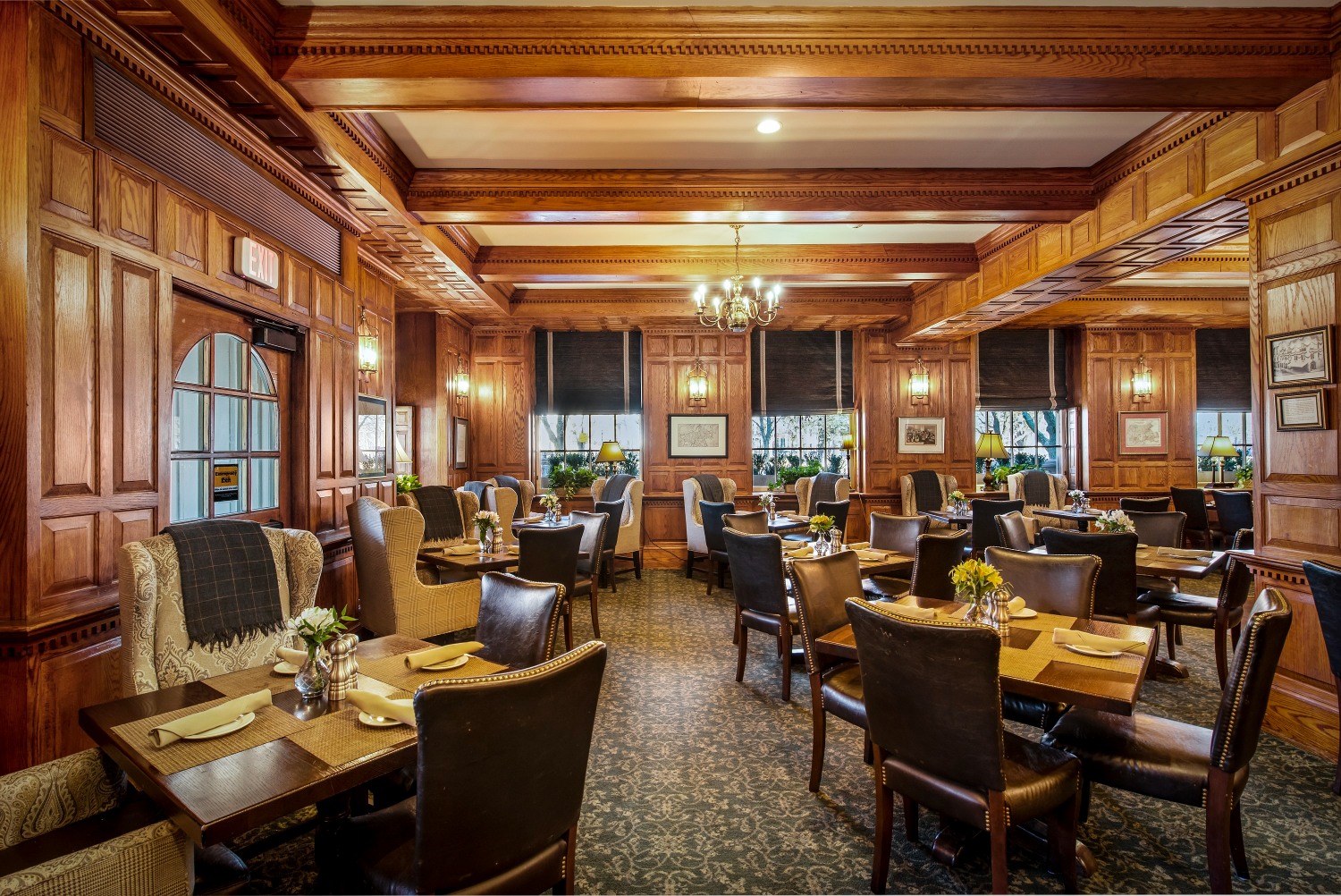 Warmly lit, upscale dining room featuring rich wooden wall paneling and coffered ceilings, dark leather dining chairs, wingback armchairs, and small tables set with fresh flowers.