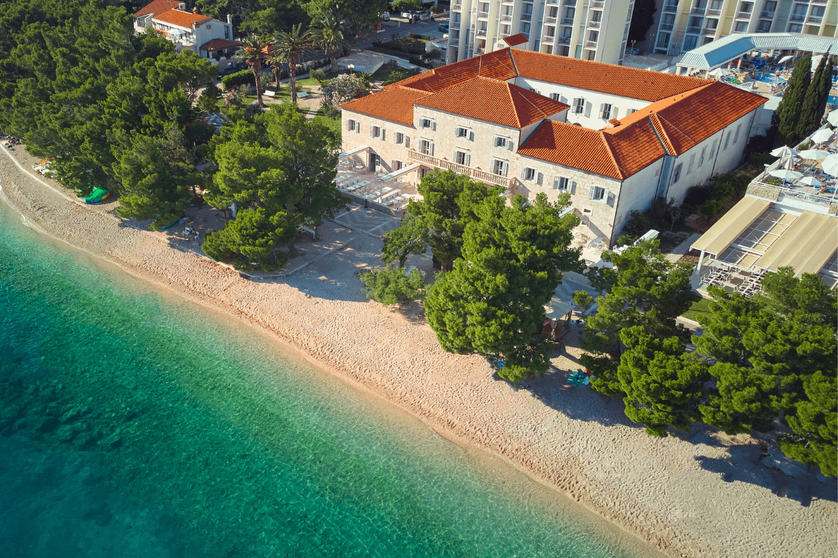 An aerial high-angle view of a coastal resort featuring a large stone building with orange tile roofs situated right along a pebble beach next to clear turquoise water, surrounded by lush green pine trees.