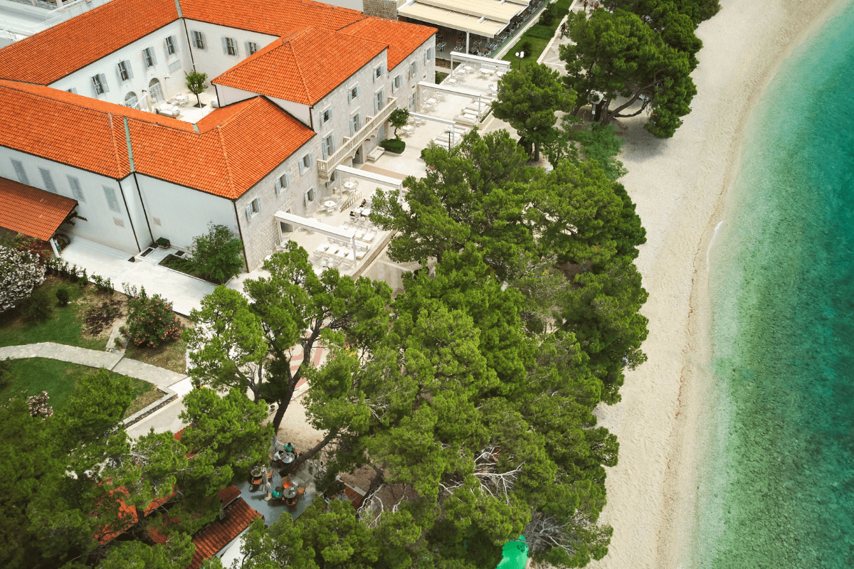 An aerial overview of a coastal hotel with orange tiled roofs, an inner courtyard, and outdoor patio seating, situated alongside a pebble beach and vibrant turquoise sea bordered by pine trees.