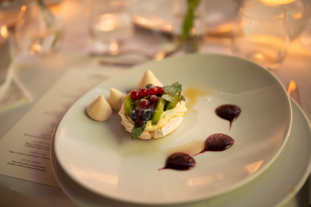 A delicately plated mini Pavlova dessert topped with fresh berries, sliced kiwi, cream, and mint leaves, accompanied by small meringue kisses and decorative berry coulis droplets on a light-colored plate, illuminated by soft candle lighting.