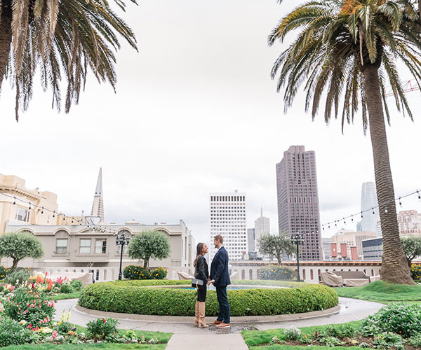 a man and woman holding hands in a park with palm trees and buildings