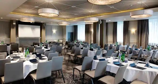 A modern, spacious conference or event hall set up with round tables draped in white tablecloths, surrounded by light grey padded chairs. The room features a projector screen at the front, stylish large circular pendant light fixtures hanging from a geome