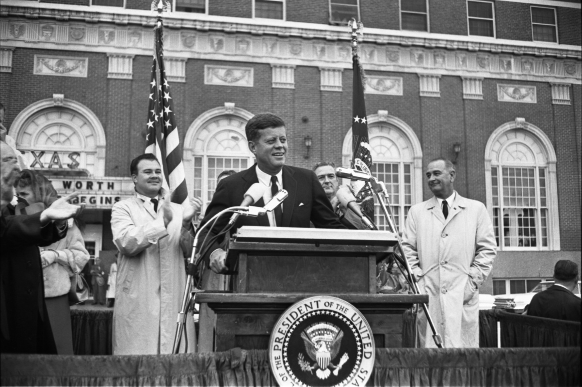 a man standing at a podium with microphones and a group of men standing behind him