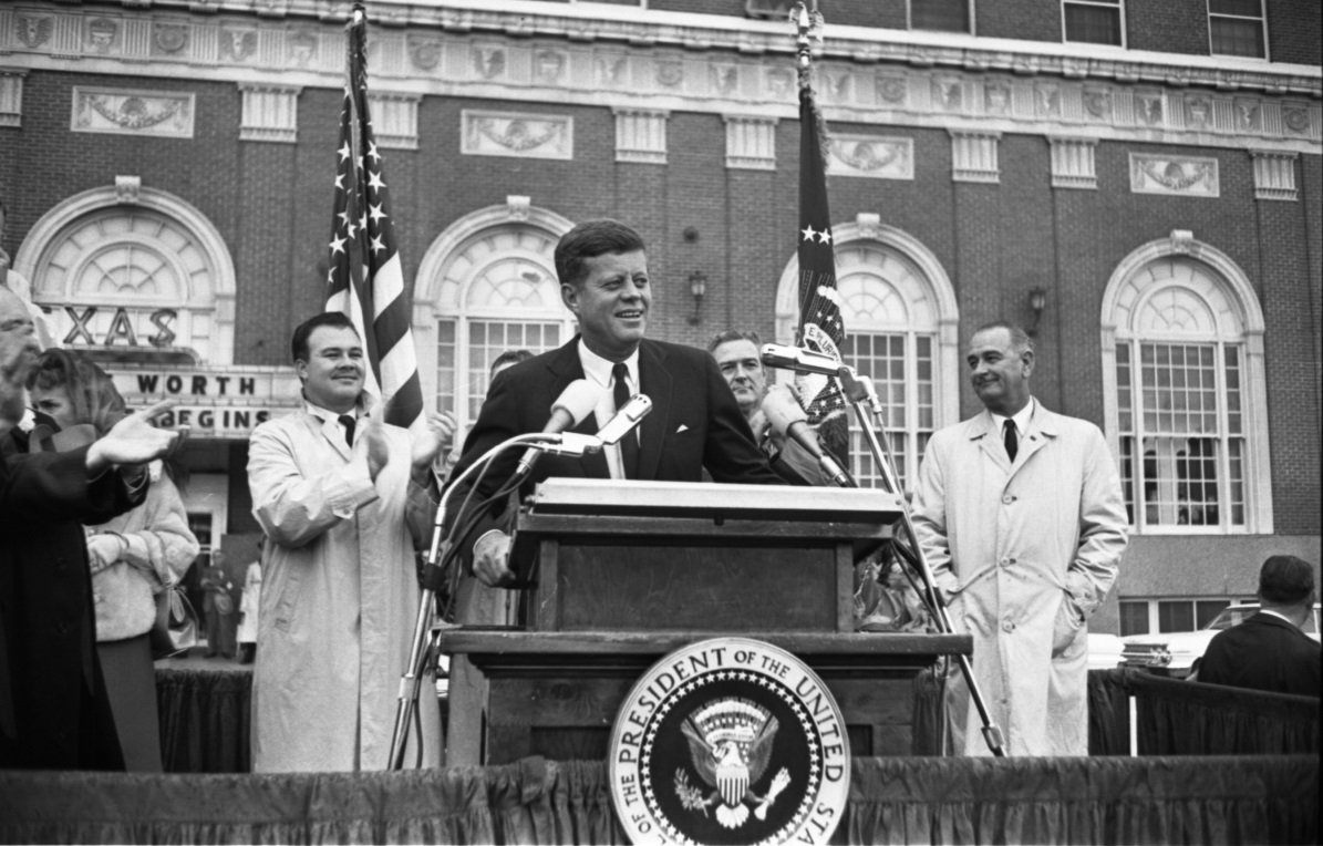 a man standing at a podium with microphones and a group of men standing behind him