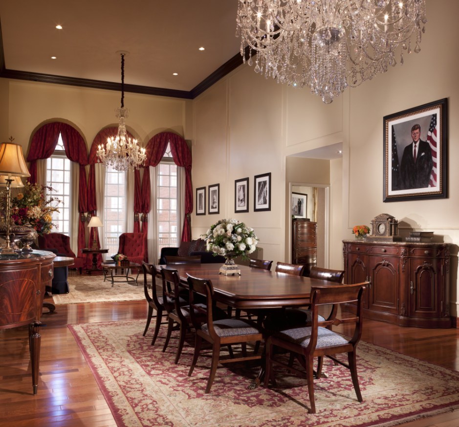Elegant hotel suite dining room featuring polished hardwood floors, a long mahogany dining table over a patterned rug, crystal chandeliers, red velvet window drapery, and a framed portrait of John F. Kennedy.